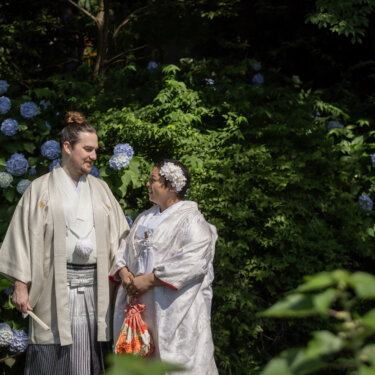 Bride and groom in white kimono smiling at each other in front of blooming blue hydrangeas, an early summer highlight of their kimono wedding photos in Japan
