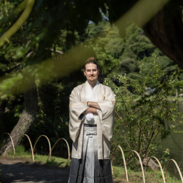 Groom in hakama standing with arms crossed beneath overhanging branches by the pond at Sankeien Garden, a solo portrait from a Yokohama pre-wedding photoshoot