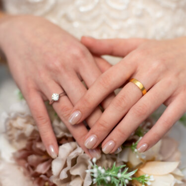 Close-up of the couple's hands layered together to show the engagement ring and gold wedding band over a dusty-pink bouquet, a classic ring detail for wedding photos.