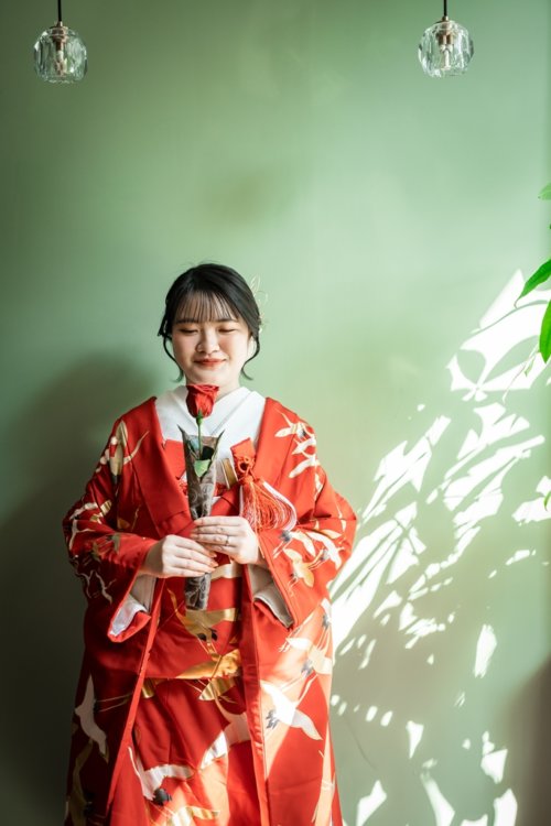 Bride smiling softly in a red crane uchikake against a sage green wall dappled with leaf shadows — kimono wedding photos in Japan