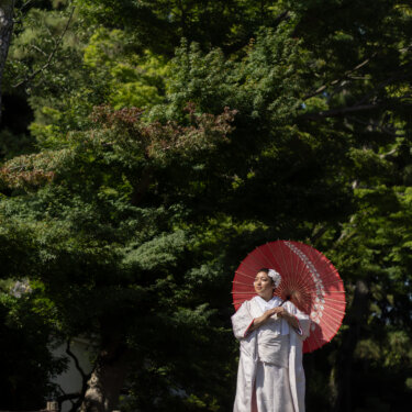 Bride in a shiromuku holding a red wagasa umbrella under deep green trees, a striking color contrast for wedding photography in a Japanese garden