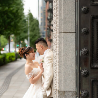 Bride and groom smiling at each other beside a bronze-studded door on a tree-lined Marunouchi street, a natural candid moment from their wedding photoshoot.