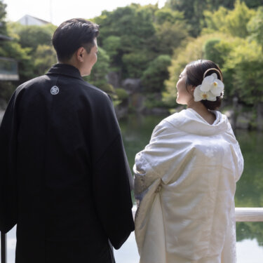 Bride and groom in traditional wedding kimono seen from behind as they look out over a garden pond, the bride wearing a large orchid hair ornament