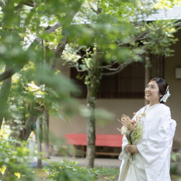 Bride in a white shiromuku holding a dried flower bouquet and laughing in front of a wooden teahouse framed by fresh green leaves