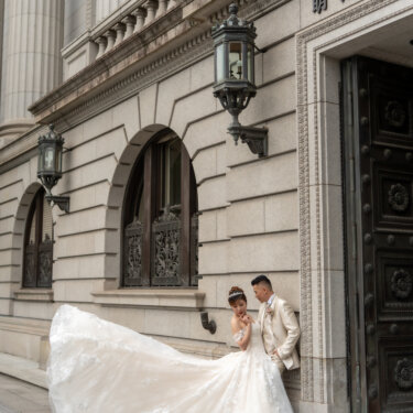 Bride sitting on the stone steps beside a bronze door as her lace train is tossed into the air and the groom leans nearby, a dynamic pose for Japan wedding photography.