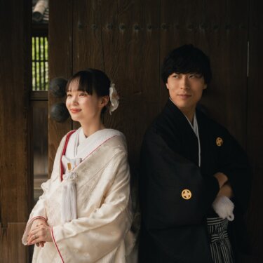 Bride in shiromuku and groom with folded arms standing back to back in front of a historic wooden gate at Sankeien Garden