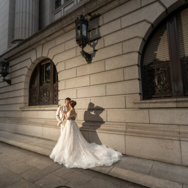 Bride and groom sharing a close moment against the granite wall of a historic Marunouchi building, their shadow cast beside iron-grilled windows and lanterns in this wedding photography shot.