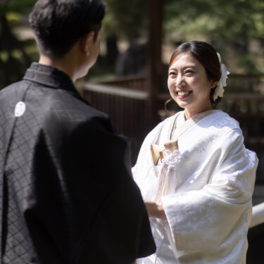 Bride in shiromuku smiling brightly at her groom, seen over his shoulder in a sunlit Japanese garden, natural moment from a kimono photoshoot