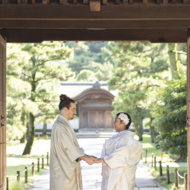Couple in shiromuku and hakama facing each other and holding hands under the frame of a wooden gate, a symmetrical composition for kimono wedding photos