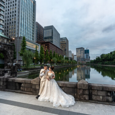 Couple embracing along the stone railing of the Imperial Palace moat, with office towers and willow trees reflected in the calm water, a daytime Marunouchi wedding photo.