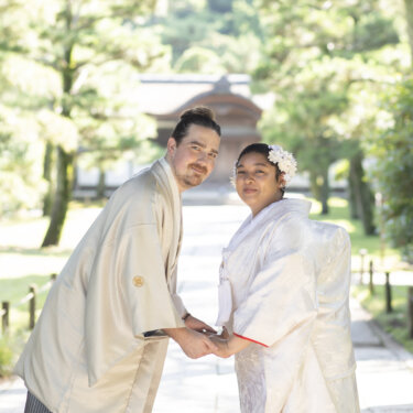 Bride and groom in white kimono holding hands and smiling at the camera through a large wooden gate at Sankeien Garden, a fun pose for pre-wedding photos in Japan