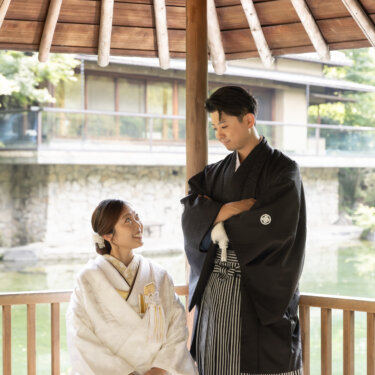 Bride seated in a garden pavilion looking up at her groom in montsuki, the pond and traditional building visible behind them