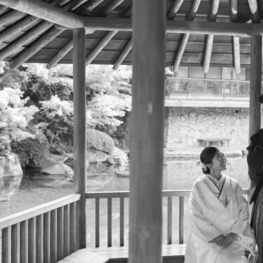 Black and white photo of a couple in wedding kimono chatting in a log pavilion above a garden pond, a relaxed candid moment