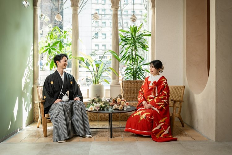 Groom in black montsuki and bride in a red crane uchikake seated facing each other among columns and greenery at our Yokohama studio — pre-wedding photoshoot in Japan