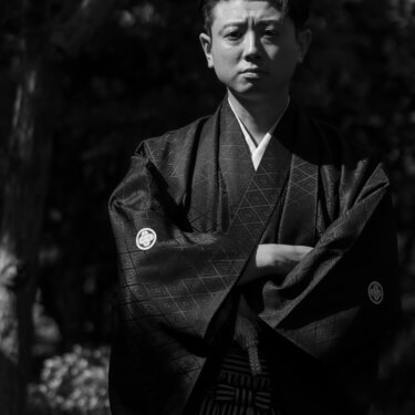 Dramatic black and white portrait of a groom in a diamond-patterned montsuki haori and striped hakama with arms crossed in dappled light