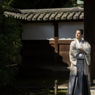 Groom in a cream kimono and striped hakama standing proudly in front of a tiled-roof gate in the shade, a solo shot from his pre-wedding photoshoot in Japan