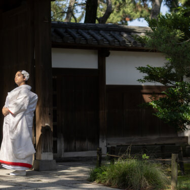 Bride in a shiromuku with a red hem looking up beside a wooden gate and white plaster wall, a solo bridal portrait from a Japan wedding photography session
