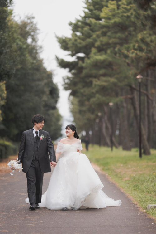 Bride and groom walking hand in hand along a pine-lined park path, a black pinstripe tuxedo paired with an off-shoulder ball gown for Japan wedding photography (TP08).