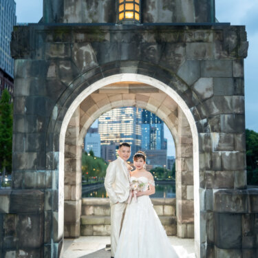 Bride and groom standing side by side in the arched stone gatehouse by the moat at twilight, a symmetrical frame for pre-wedding photoshoot ideas in Tokyo.