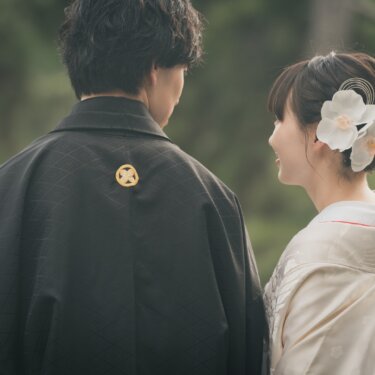 Back view of a groom in montsuki with a gold family crest beside a bride with an orchid hair ornament turning to smile at him