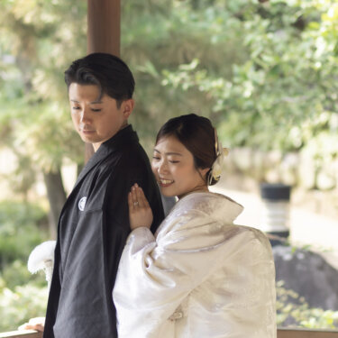Bride in a crane-patterned shiromuku resting her hand on her groom's back in a wooden garden pavilion, a tender moment from a kimono wedding photoshoot