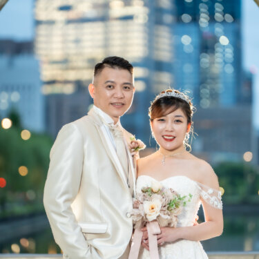 Smiling couple in a champagne tuxedo and off-shoulder gown with a tiara, holding a dusty-pink bouquet with the Tokyo skyline softly blurred behind them, from their pre-wedding photoshoot.