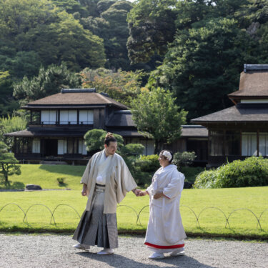 Groom leading the bride by the hand across the lawn in front of Rinshunkaku at Sankeien Garden, a natural walking shot from their kimono wedding photos in Yokohama