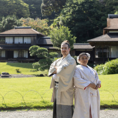 Couple in white kimono posing back to back with arms crossed on a sunny garden lawn, with sukiya-style buildings and pine trees behind them in this pre-wedding photoshoot