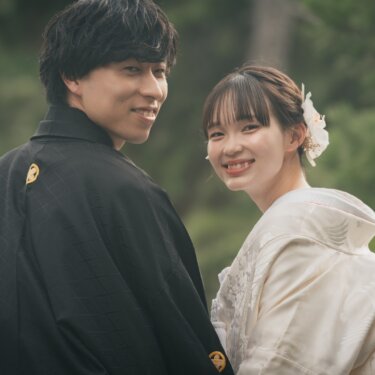 Bride in shiromuku and groom in black montsuki smiling over their shoulders against deep green trees as part of their Japan wedding photography session