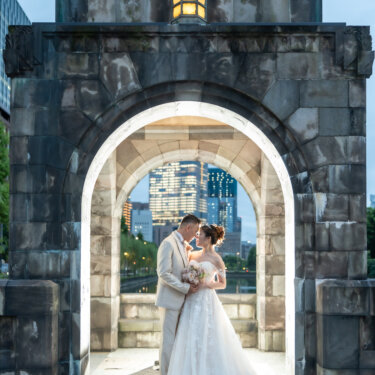Couple leaning in forehead to forehead under a lit stone arch with Marunouchi skyscrapers seen through the opening, a romantic kiss-close pose for Tokyo wedding photography.