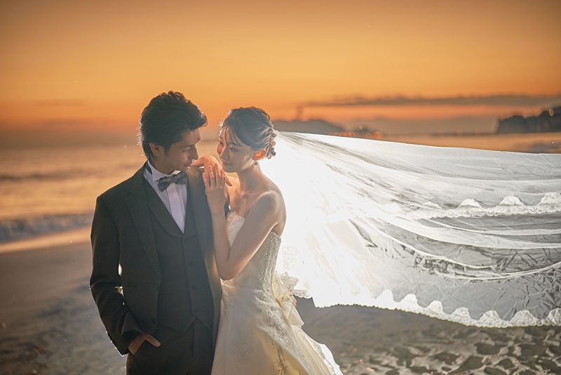 The bride's veil billows dramatically in the sea breeze as the couple embraces at sunset.