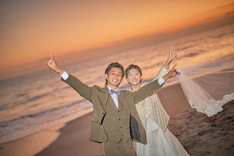 Groom in an olive three-piece suit and bride in a flowing cream gown raise their arms in celebration on the beach at golden-hour sunset.
