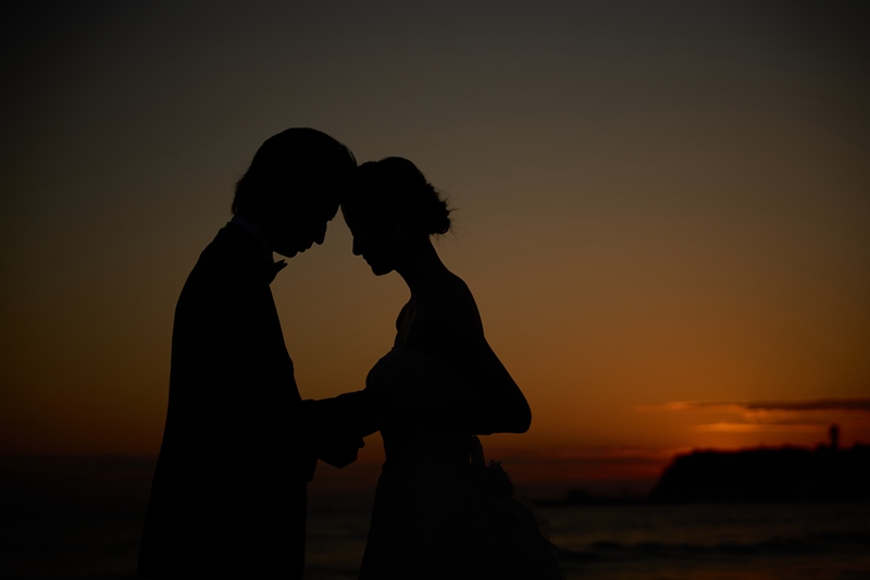 A silhouetted couple embracing forehead to forehead on the beach at Shichirigahama, a lighthouse visible on the point at sunset.