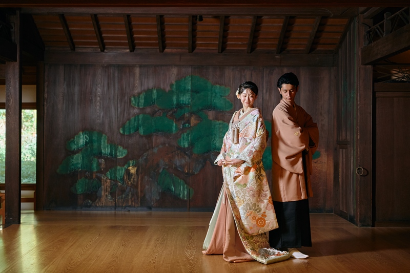 Newlyweds in richly patterned kimono standing before a hand-painted pine screen inside a traditional Japanese room.