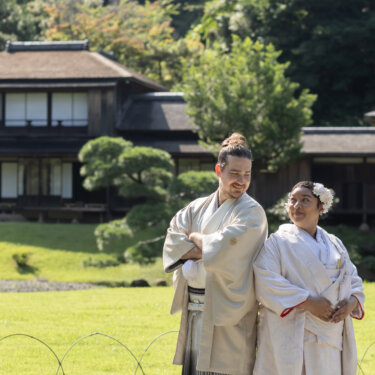 Groom and bride standing back to back on the lawn in front of a traditional villa at Sankeien Garden, a playful pose for Japan wedding photography in white kimono