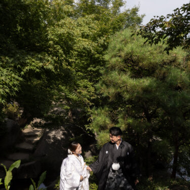 Couple in traditional wedding kimono holding hands beside a garden pond framed by pine and maple trees, a serene pre-wedding photoshoot in Japan