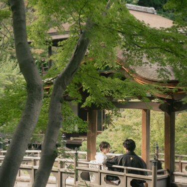 Couple in kimono sitting on a roofed wooden bridge framed by fresh green maple trees at Sankeien Garden, Yokohama