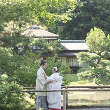 Bride in shiromuku and groom in hakama embracing beside a pond under green maple and pine trees, a vertical frame from kimono wedding photos in a Japanese garden