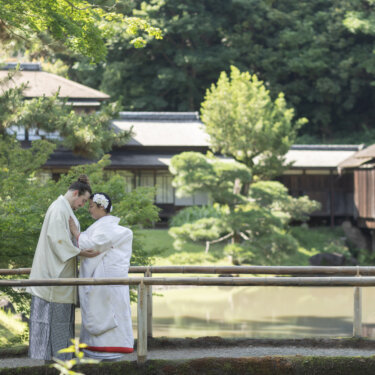 Couple in shiromuku and hakama touching foreheads by the pond with the historic Rinshunkaku villa behind them, a romantic pose from their Sankeien Garden wedding photoshoot