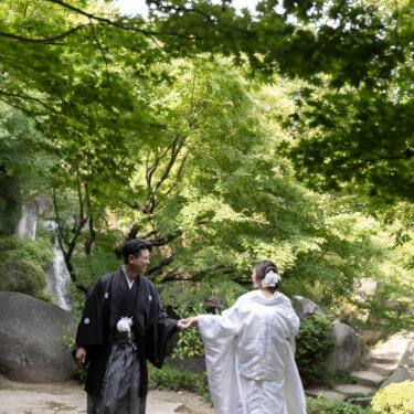 Groom in montsuki leading his bride in shiromuku by the hand along a stone path under fresh green maples, with a small waterfall behind them in a Japanese garden