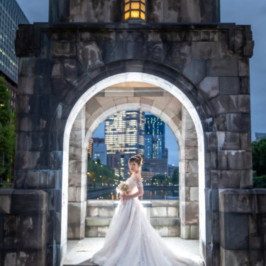 Bride standing alone inside the glowing stone archway beside the Imperial Palace moat at dusk, her long lace train trailing down the steps in this Japan wedding photography shot.