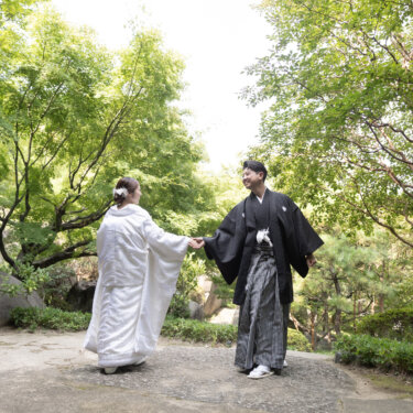 Bride in shiromuku reaching back for her groom in montsuki on a stone path beside a waterfall, surrounded by fresh green maples in a Japanese garden