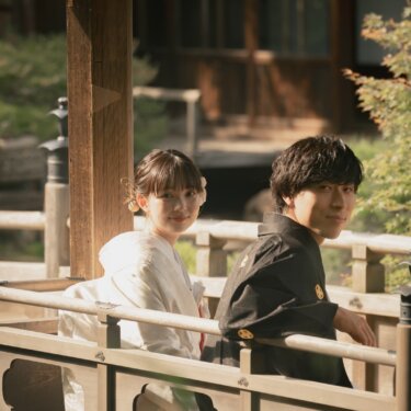 Couple in white shiromuku and black montsuki looking back from a roofed wooden pavilion bridge in the Japanese garden of Sankeien