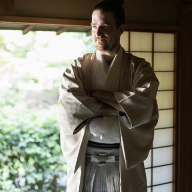 Groom in a cream haori and striped hakama standing with arms crossed by the shoji screens of a traditional house at Sankeien Garden for his kimono wedding portrait