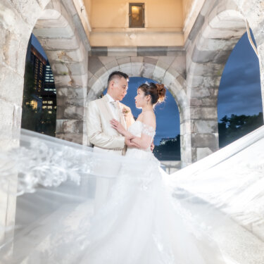 Bride and groom embracing beneath a stone arch while her flowing veil sweeps across the frame, a dramatic motion shot for pre-wedding photos in Japan.