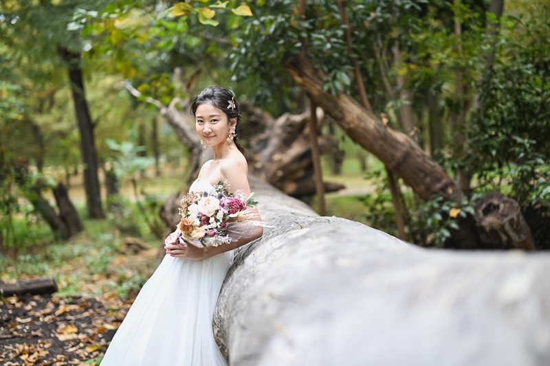 Bride resting against a fallen mossy log, holding a bouquet of dried flowers in a wooded corner of Osen Ryokuchi Park.
