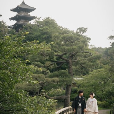 Couple in kimono walking across a wooden bridge with Sankeien Garden's three-story pagoda rising above the pines in Yokohama