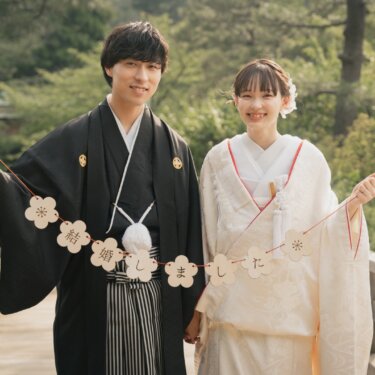 Couple in shiromuku and montsuki holding a flower-shaped garland reading 'we got married' in Japanese on a wooden bridge at Sankeien Garden