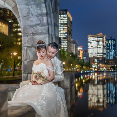Groom hugging his seated bride from behind under a stone arch as the city lights reflect on the moat at blue hour, a soft and intimate wedding photo pose.