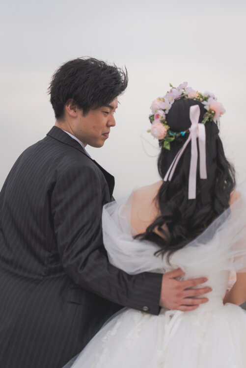 Back view of the groom holding his bride by the waist as he gazes at her flower crown and ribbon, a pre-wedding photoshoot in Japan in the Black Large tuxedo (TP08).